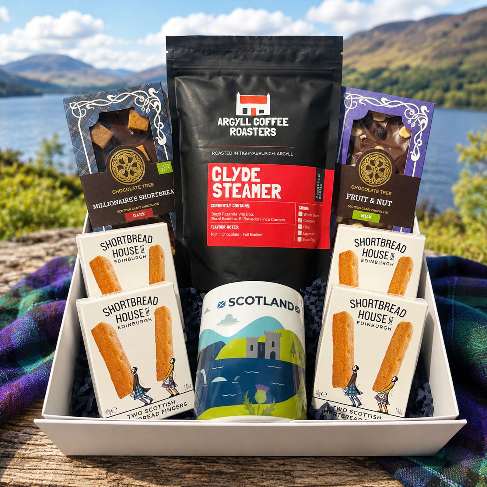 A gift basket filled with Scottish snacks and treats, including Argyll Coffee Roasters Clyde Steamer, Shortbread House English Treats, and a Scotland-themed mug, set outdoors on a wooden surface with a plaid blanket and a scenic lake and mountains in the background.