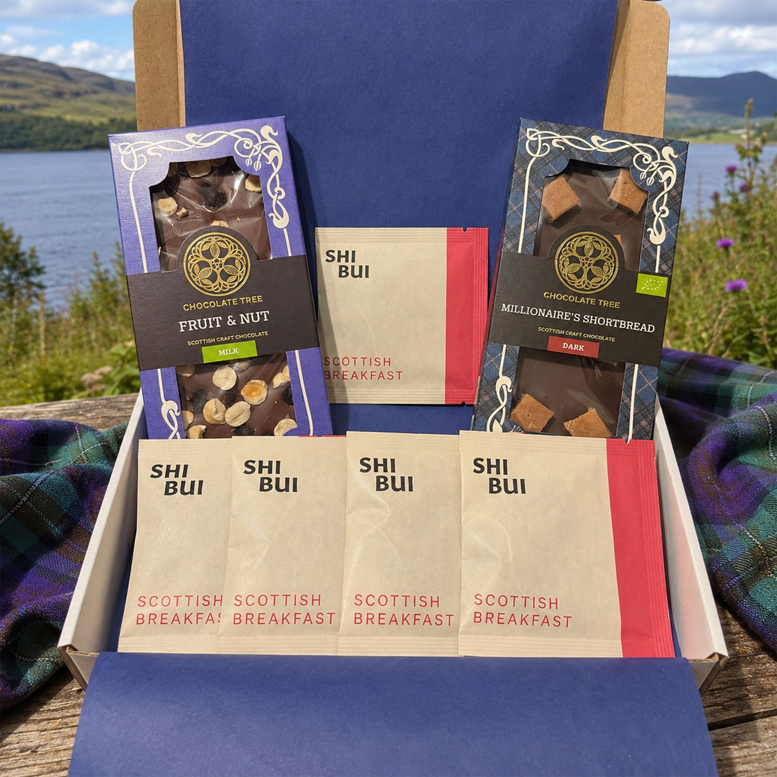A gift box of Scottish breakfast treats is displayed outdoors on a wooden surface with a scenic loch and mountains in the background. The open box contains two packaged chocolate bars, three single-serve packets, and a small packet of Scottish Breakfast Cereal.